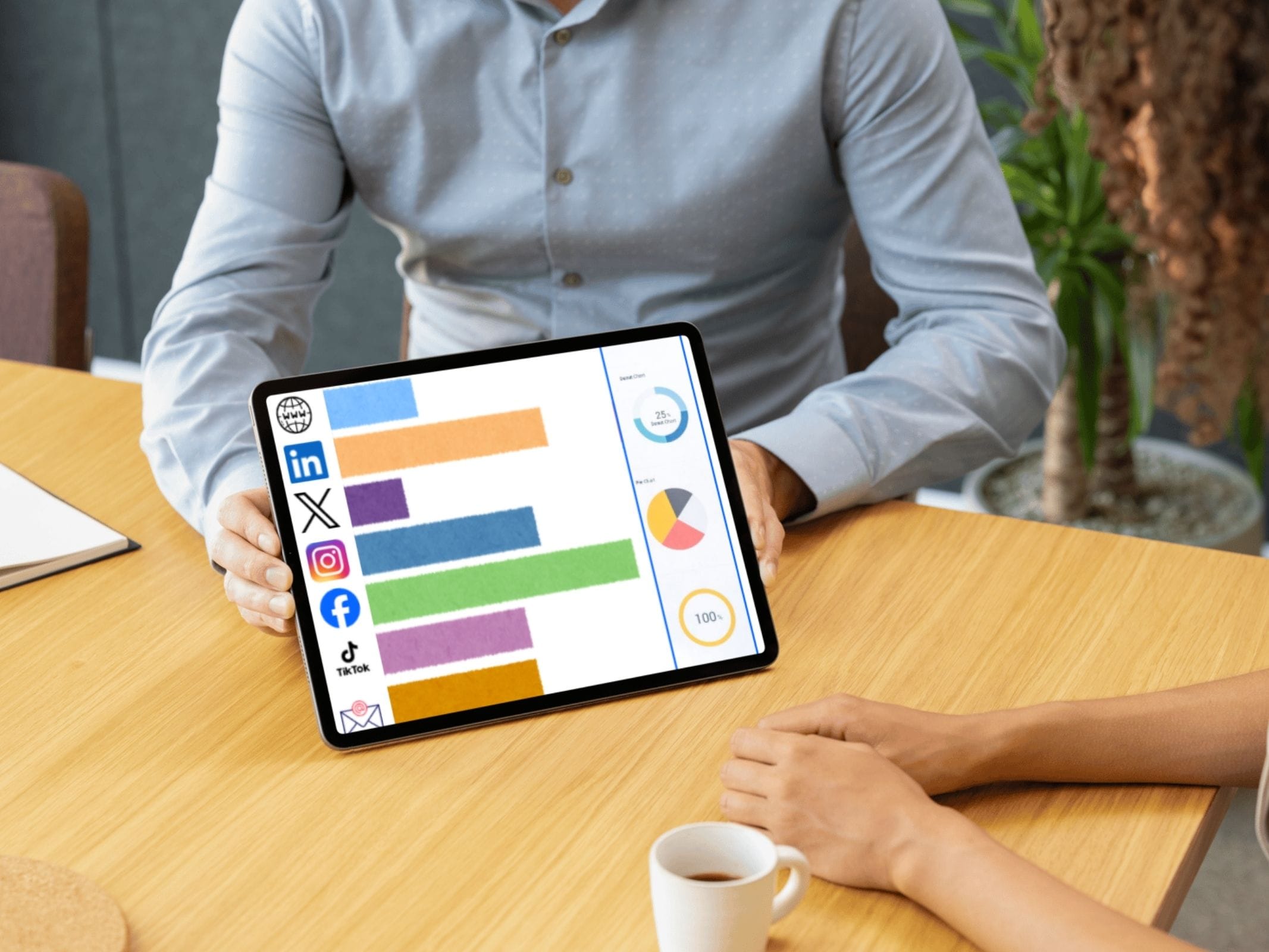 Zwei Personen bei einem Gespräch an einem Tisch bei dem eine Person frontal ein Tablet mit einem Balkendiagramm zu verschiedenen Medienkanälen zeigt während die andere Person im Seitenprofil mit einer Kaffeetasse daneben sitzt.